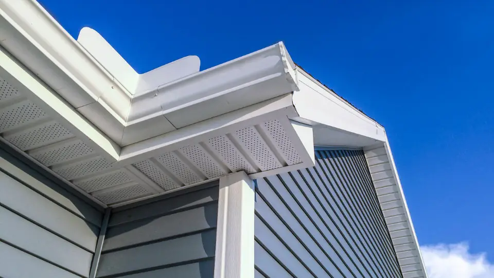 New white seamless gutter installed on the corner of a residential home with vinyl siding, under a clear blue sky.